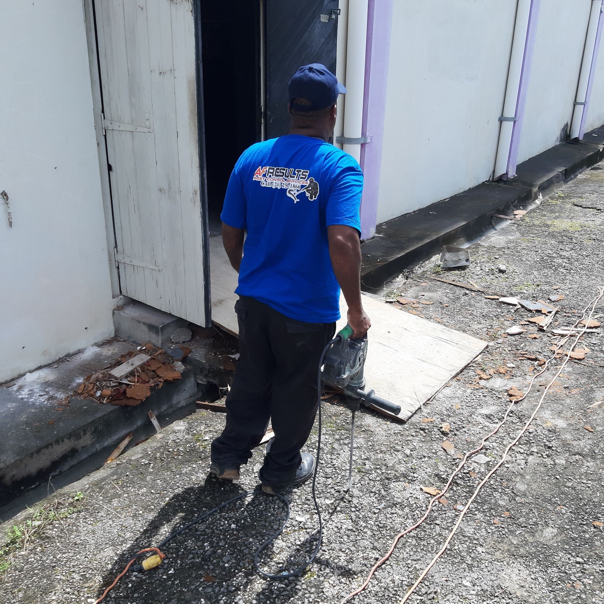 Worker in a blue shirt holding a power tool, standing outside a building beside an open door.